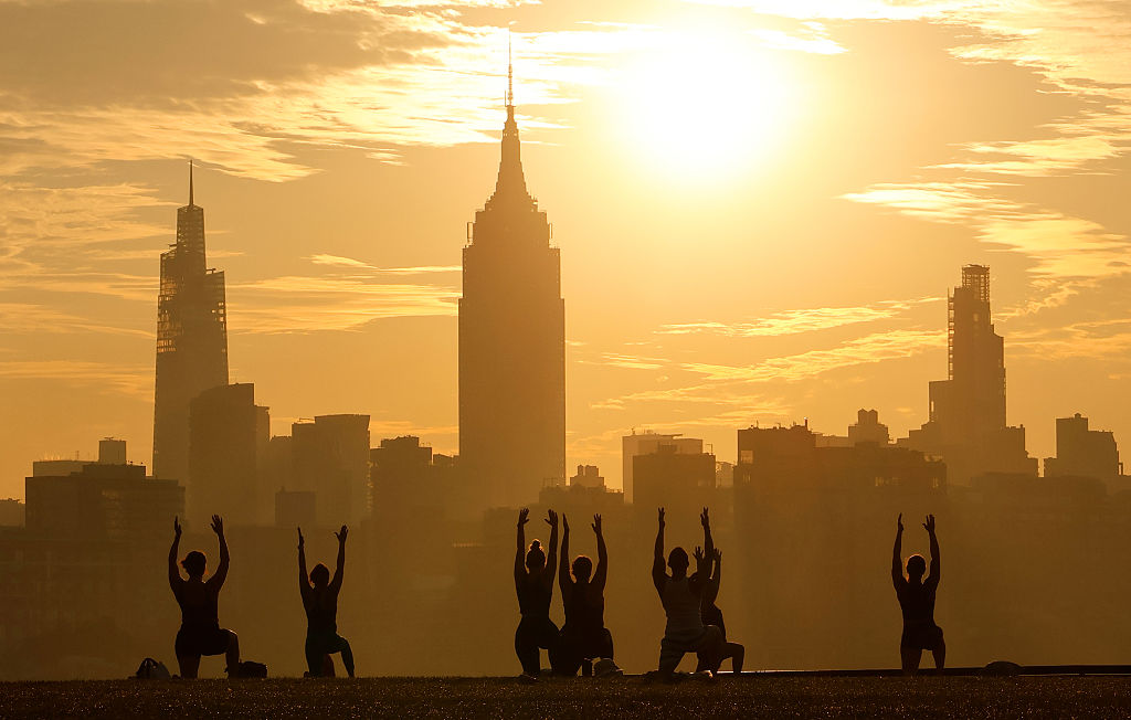 HOBOKEN, NJ - JULY 29: The sun rises behind the skyline of midtown Manhattan and the Empire State Building in New York City as people participate in a yoga class next to the Hudson River on July 29, 2025, in Hoboken, New Jersey.
