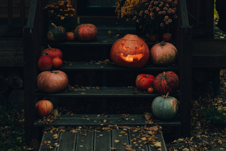 Spooky carved pumpkin illuminating porch steps decorated with various pumpkins, flowers and autumn leaves, creating a festive halloween scene