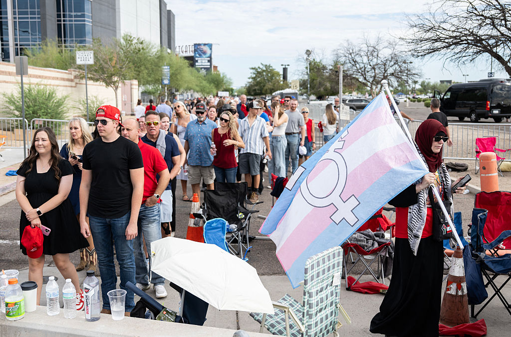 A protester carrying a transgender support flag walks past people waiting to attend the memorial for Charlie Kirk at State Farm Stadium in Glendale, Arizona, on September 21, 2025. (Photo by ANDREW CABALLERO-REYNOLDS / AFP) (Photo by ANDREW CABALLERO-REYNOLDS/AFP via Getty Images)